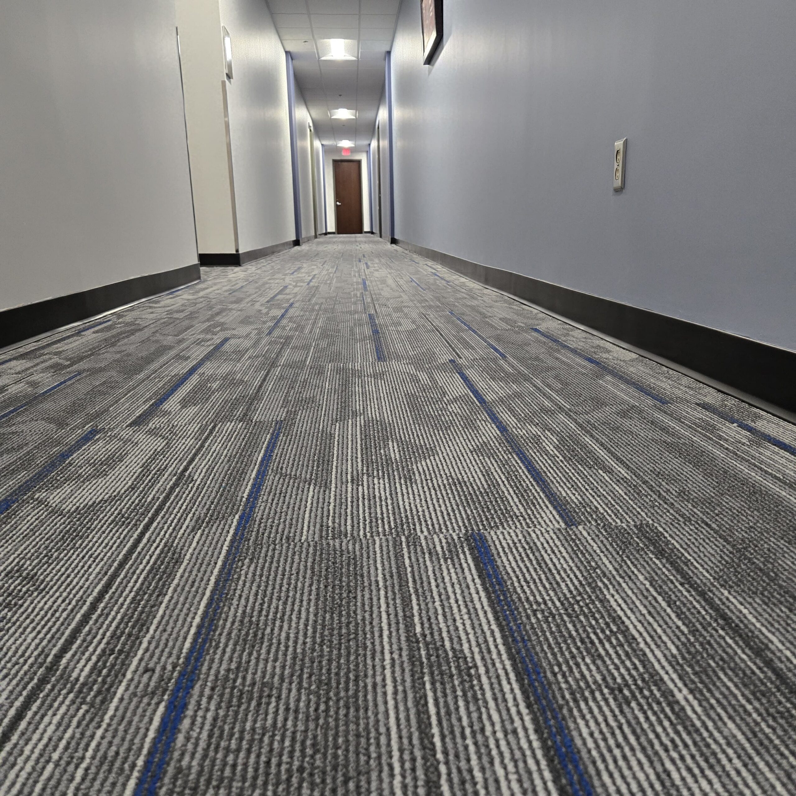 A low-angle view of a long, empty hallway with blue and gray carpet, light blue walls, ceiling lights, and a closed door at the end.