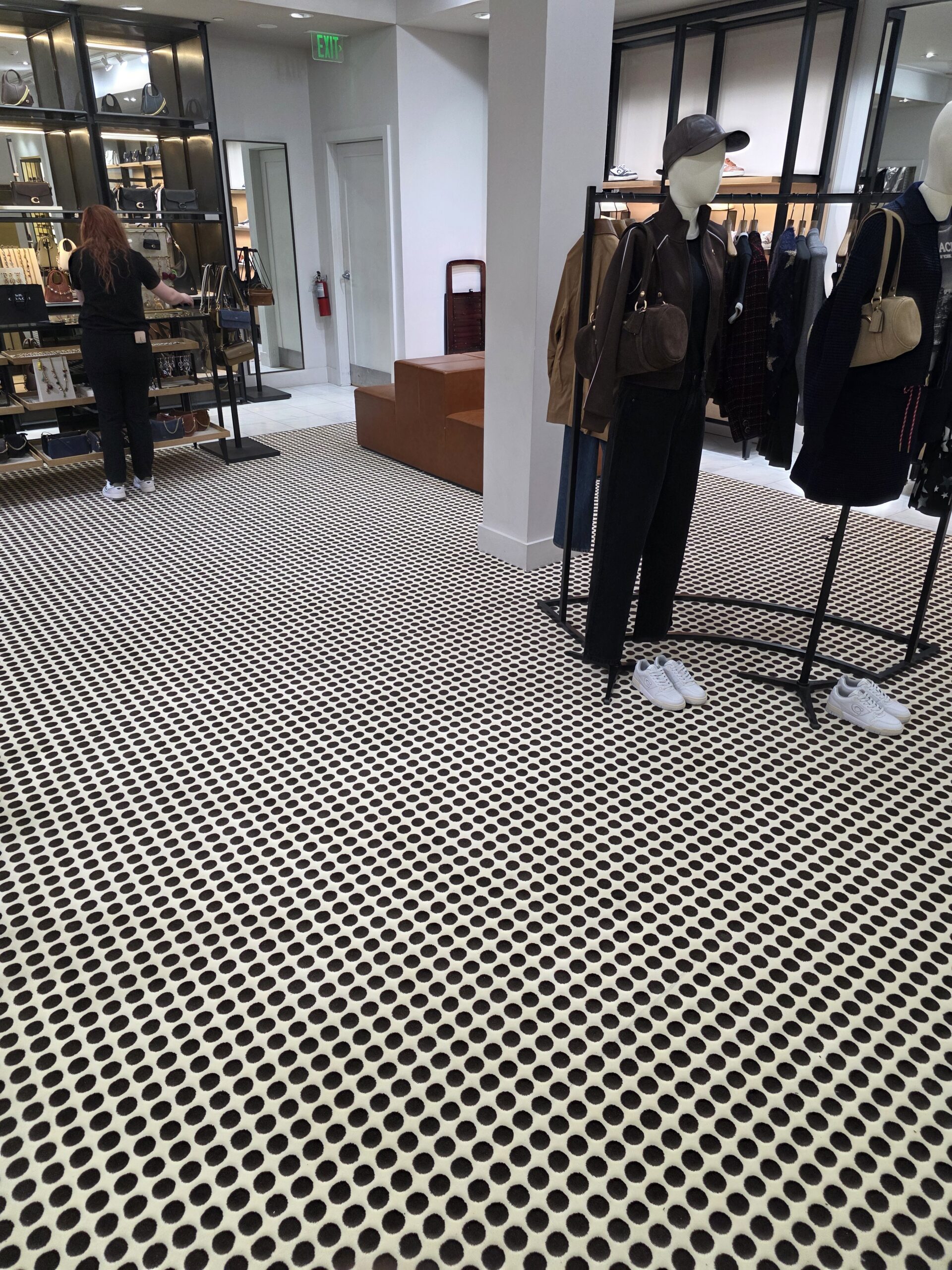 Clothing store interior with mannequins displaying outfits, a rack of clothes, and a patterned floor with black dots. A person is browsing shelves in the background.