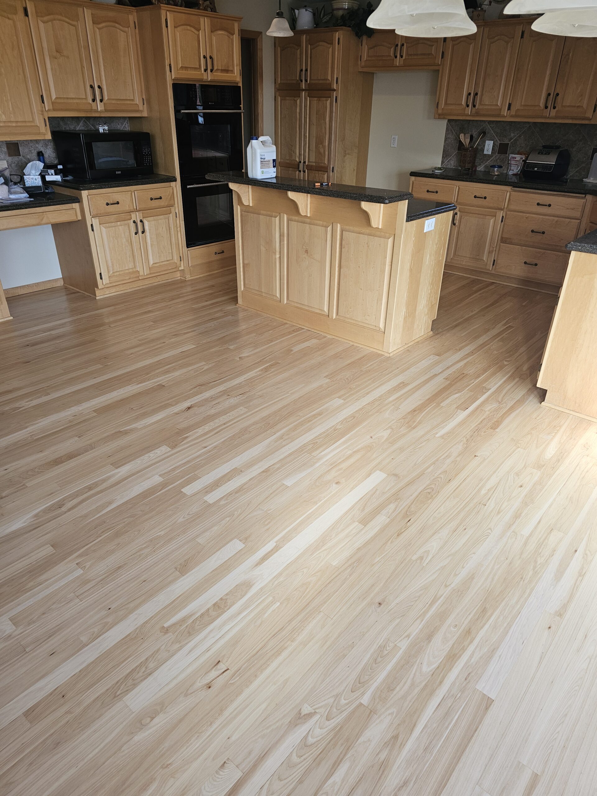 A spacious kitchen with light wood cabinets, granite countertops, and newly installed light wood flooring. An island stands at the center with two pendant lights overhead.