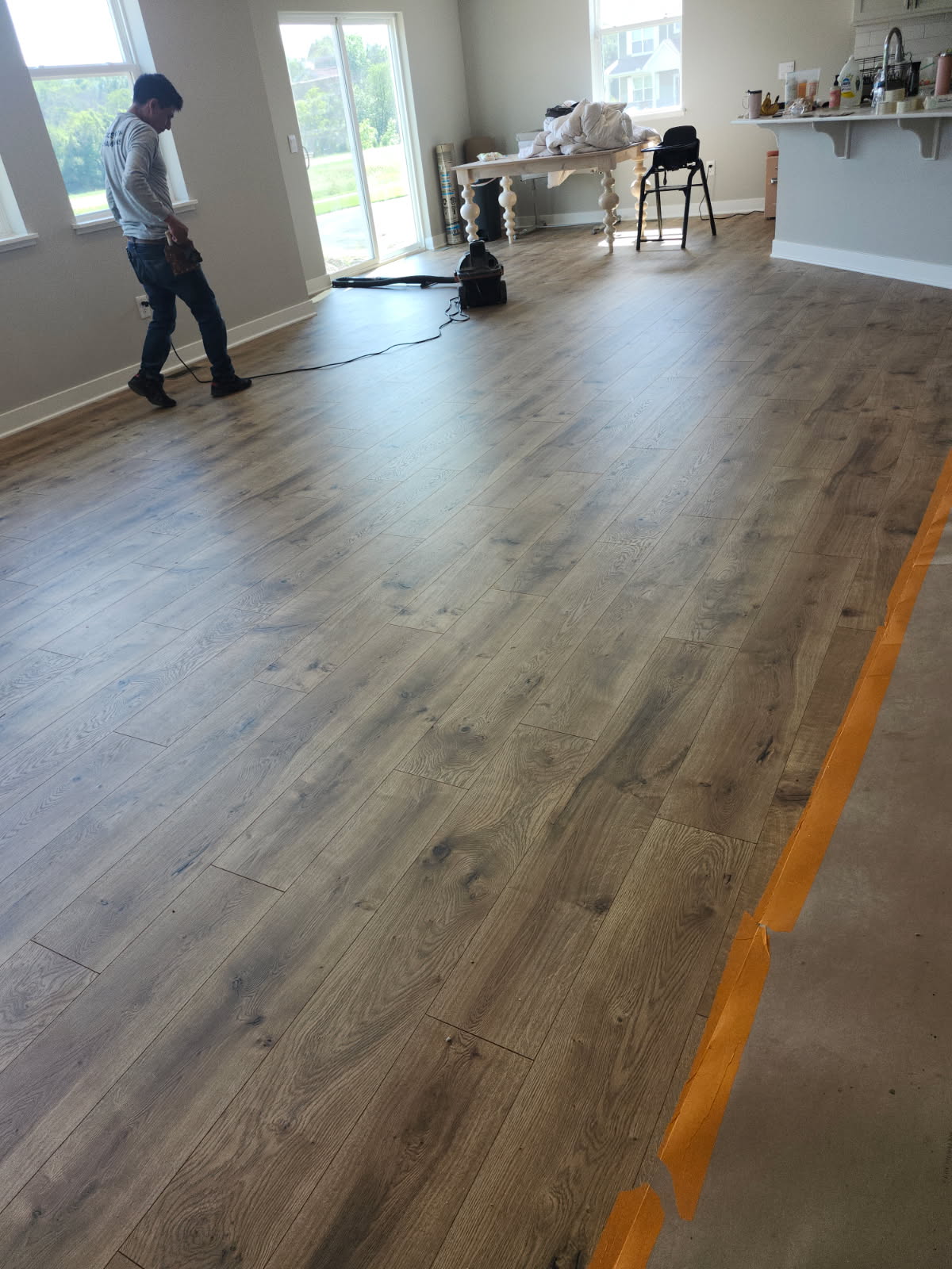 A person stands in a large room with newly installed wood flooring; tools and supplies are visible, and an orange tape line marks the floor.
