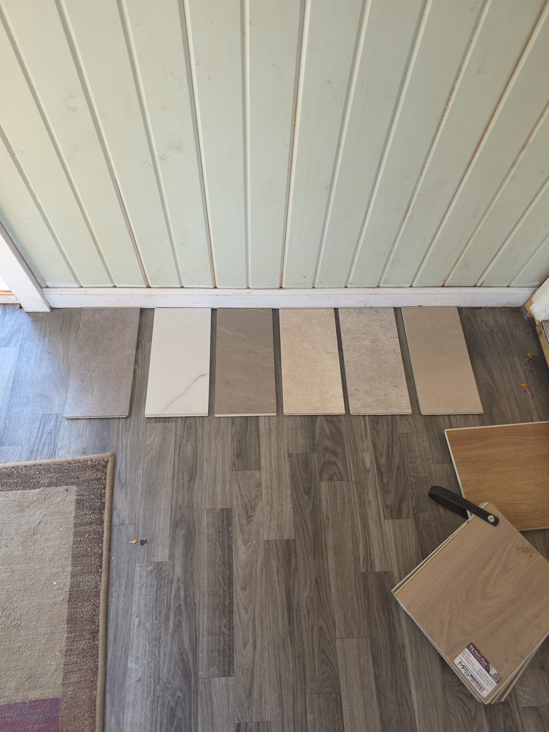 Six rectangular floor tile samples are arranged side by side on gray wood-patterned flooring near a light green paneled wall.