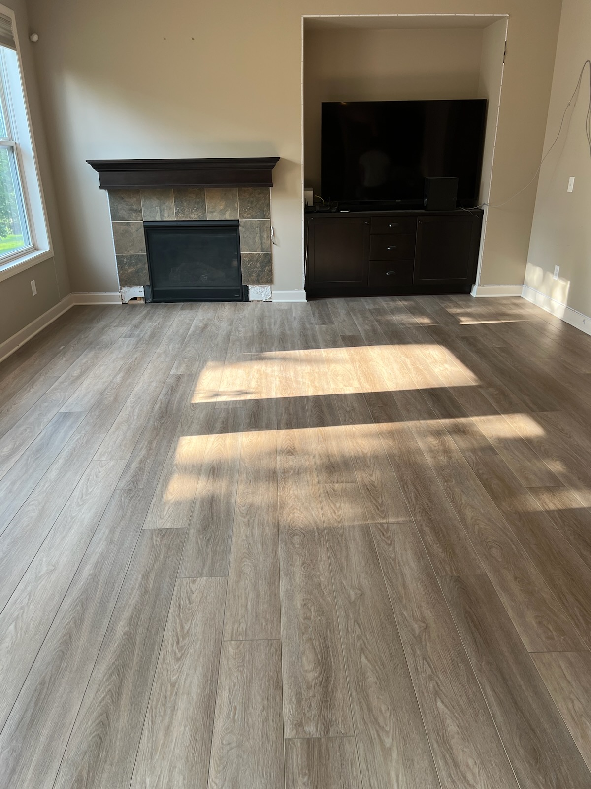 A living room with wood-look flooring, a fireplace with a dark mantel, a built-in TV cabinet, and sunlight streaming in from a window on the left.